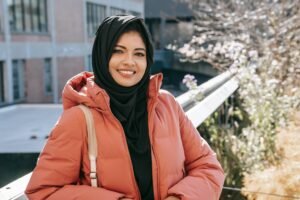 Cheerful ethnic female in hijab and jacket smiling and looking at camera on street